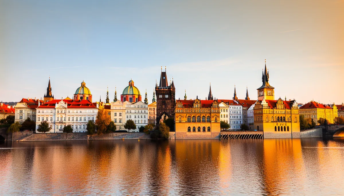 Beautiful sunset view of Prague's historic city center with traditional red-tiled rooftops, church spires, and the Vltava River reflecting warm golden light