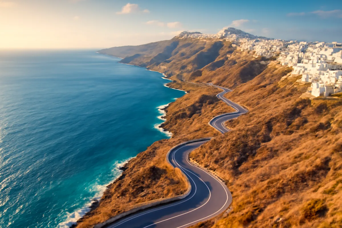 Aerial view of a scenic Greek coastal road winding along dramatic cliffs above the turquoise Aegean Sea, with whitewashed hilltop villages bathed in golden hour light