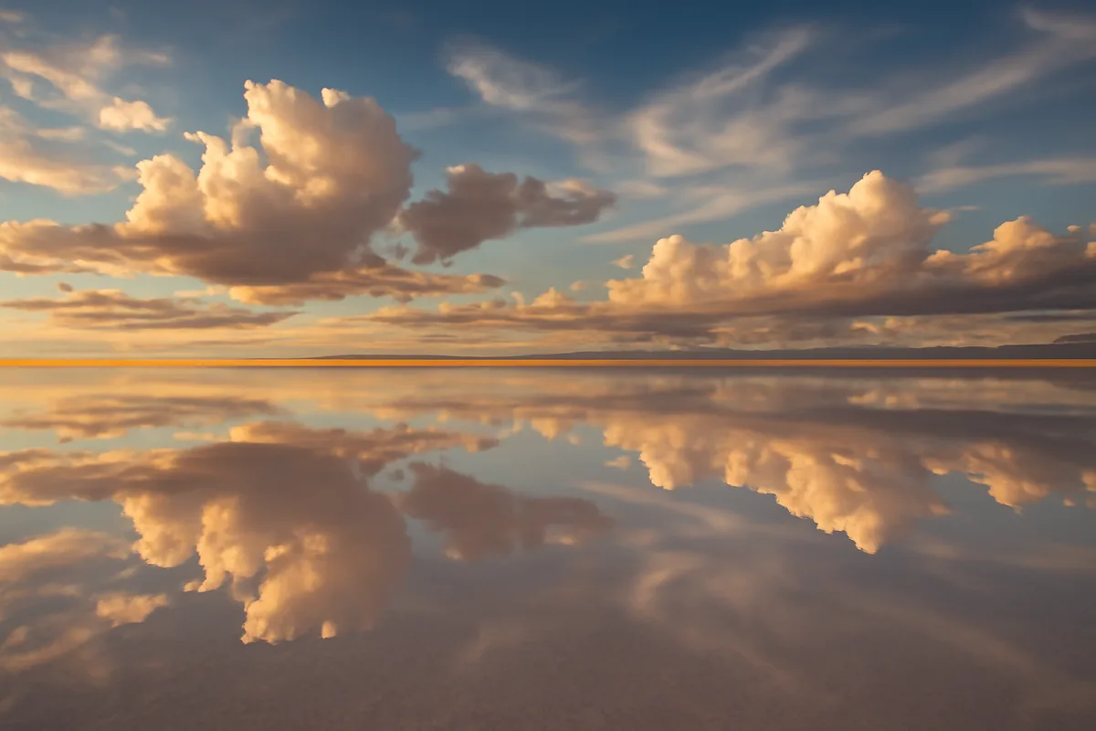 The vast white expanse of Bolivia's Salar de Uyuni salt flats reflecting a dramatic golden sky at sunset, creating a perfect mirror image on the horizon