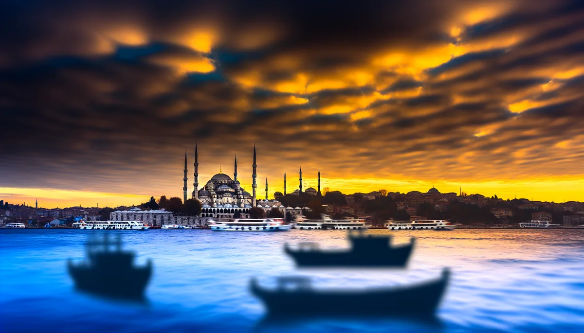 Stunning sunset view of Istanbul's Blue Mosque with its distinctive minarets silhouetted against a dramatic sky, with the Bosphorus strait and traditional boats in the foreground