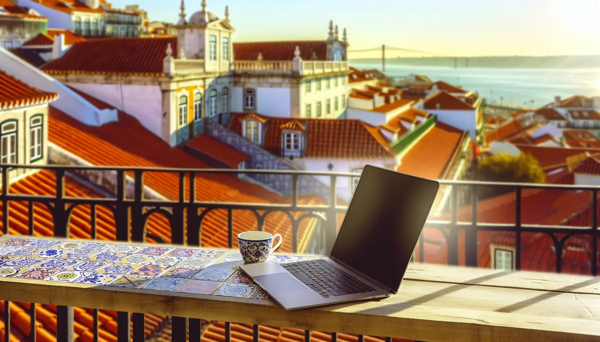 Scenic outdoor workspace terrace overlooking Lisbon's traditional rooftops and river, with laptop and coffee setup in golden hour lighting