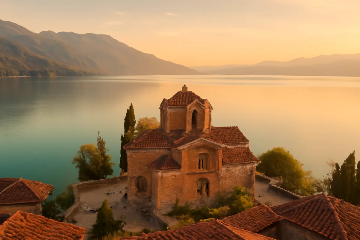 A historic Byzantine church sitting on the tranquil shores of Lake Ohrid in North Macedonia, surrounded by mountains bathed in golden evening light