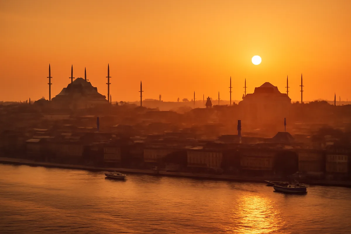 Aerial panoramic view of Istanbul's iconic skyline at golden hour, with the Bosphorus strait reflecting warm orange light and historic mosques and minarets rising against a glowing amber sky.
