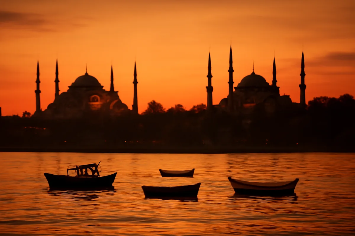 Panoramic golden hour view of Istanbul's iconic skyline featuring the Hagia Sophia and Blue Mosque domes reflected in the calm Bosphorus strait, with traditional wooden boats in the foreground.